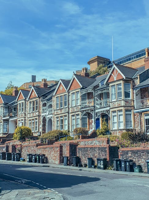 A row of Victorian-style terraced houses with ornate ironwork balconies and decorative gables, situated along a paved street with a brick retaining wall and several black wheelie bins lined up at the curb. The houses feature large bay windows, some with white frames, and brick facades mixed with light-colored plaster finishes. Behind the row, modern office or commercial buildings are visible, partially obscured by trees and the rooftops. The sky above is clear with a few wispy clouds, indicative of a bright day. The scene depicts an urban residential area where residents may arrange private waste disposal services, as evidenced by the household rubbish bins aligned outside. The photograph captures the blend of historic architecture with contemporary urban infrastructure, aligned with the context of independent waste collection and rubbish removal services such as those offered by Waste Removal Dulwich, in a setting that typically involves on-site clearance or private disposal solutions.