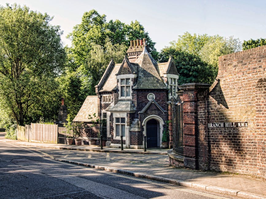 A historic Victorian-style house with intricate brickwork and decorative stone detailing is situated behind a wrought iron gate set within a brick wall, located on a quiet residential street. The building features a steeply pitched roof with multiple gables, adorned with slate tiles, and several small chimney stacks. The windows are tall with white frames, some set within bay windows that project outward, adding architectural interest. To the left, a wooden fence separates the property from the pavement, which runs alongside a tarred road with double yellow lines. Mature trees with lush green foliage surround the property, providing a natural backdrop and partial shade. Sunlight illuminates the scene, casting soft shadows on the brick wall and the pavement, suggesting a clear day. The street sign reading 'BRANCH HILL N.W.3' is affixed to the brick wall, indicating the location. This setting exemplifies a private property that might benefit from professional waste removal services, aligning with independent collection or on-site clearance options offered by Waste Removal Dulwich for period properties and flats.