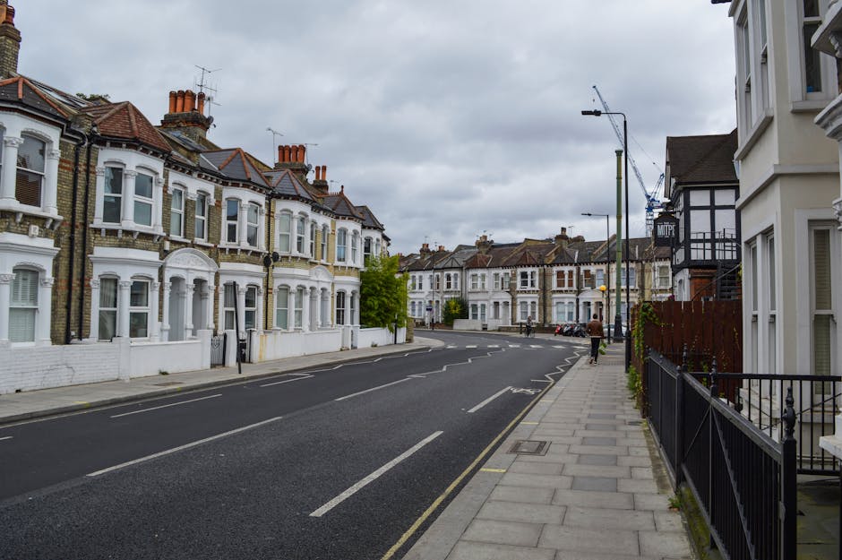 A quiet residential street lined with Victorian terraced houses featuring white facades, bay windows, and brickwork details, with some buildings having ornate architectural features. The pavement on the right side is paved with large, light grey slabs, complemented by black metal railings and small plants along the edge. On the left, there is a black asphalt road with white dashed and solid lines marking parking spaces, which are mostly empty. Several cars are parked along the far side of the street, with some visible in the distance. Tall street lamps with simple, modern designs extend over the pavement, and a construction crane can be seen in the background indicating ongoing development. The sky is overcast with grey clouds, giving the scene a muted, daytime atmosphere. The scene is typical of an area where private waste removal services, such as those offered by Waste Removal Dulwich, could handle rubbish collection from local residential properties, supported by the tidy and well-maintained environment.