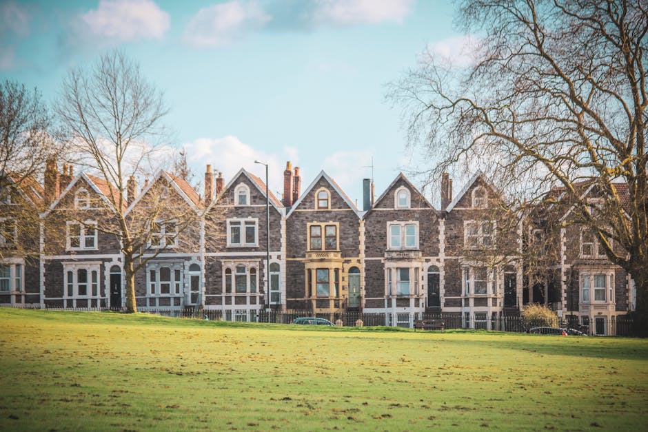 A historic Victorian-style house with intricate brickwork and decorative stone detailing is situated behind a wrought iron gate set within a brick wall, located on a quiet residential street. The building features a steeply pitched roof with multiple gables, adorned with slate tiles, and several small chimney stacks. The windows are tall with white frames, some set within bay windows that project outward, adding architectural interest. To the left, a wooden fence separates the property from the pavement, which runs alongside a tarred road with double yellow lines. Mature trees with lush green foliage surround the property, providing a natural backdrop and partial shade. Sunlight illuminates the scene, casting soft shadows on the brick wall and the pavement, suggesting a clear day. The street sign reading 'BRANCH HILL N.W.3' is affixed to the brick wall, indicating the location. This setting exemplifies a private property that might benefit from professional waste removal services, aligning with independent collection or on-site clearance options offered by Waste Removal Dulwich for period properties and flats.