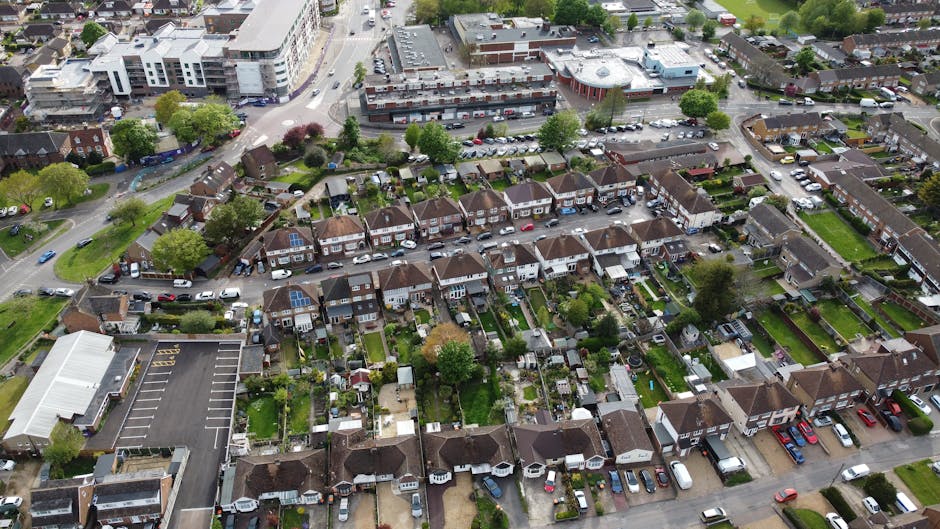 An aerial view of a suburban residential area showing a row of terraced houses with small, well-maintained gardens and driveways. The houses have brown tiled roofs, some with solar panels, and a mix of brick and painted exteriors. In the foreground, a large parking lot with marked spaces and multiple parked cars is visible adjacent to a single-story building. Surrounding the houses, there are trees, green lawns, and narrow pathways connecting the properties. The background features a busy street with additional commercial and residential blocks, including larger apartment buildings and a parking area filled with vehicles. The scene is captured in daylight with natural light illuminating the scene evenly, highlighting the residential and urban environment. The image exemplifies typical urban sprawl with a combination of private gardens and shared parking spaces, reflecting aspects of modern waste management and independent disposal practices, as often handled by local rubbish removal services, such as Waste Removal Dulwich.