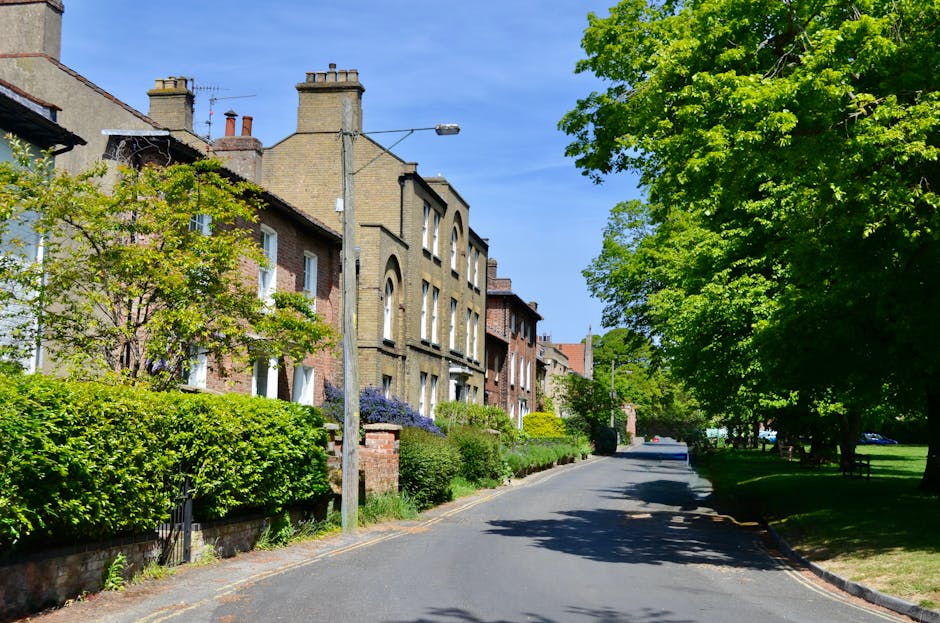 A residential street scene featuring a row of traditional brick houses with varied architectural styles, some with chimney stacks and arched window details, situated on the left side of the image. In front of the houses, there are neatly maintained gardens with green shrubs, flowering plants, and small trees, adding vibrant colors and textures to the scene. The street itself is paved with asphalt and curves gently to the right, bordered by a curb with a narrow sidewalk. On the right side, a large, leafy tree with bright green foliage provides substantial shade, its branches extending over the roadway. A lamppost is positioned near the center of the image, slightly leaning to the right, with a single streetlight attached. Above, the sky is clear and blue, suggesting a bright, sunny day. The scene exemplifies a peaceful, suburban environment that could be a typical setting for private rubbish removal or on-site clearance services by Waste Removal Dulwich, with no visible vehicles or trash, emphasizing the clean, orderly atmosphere of the area.