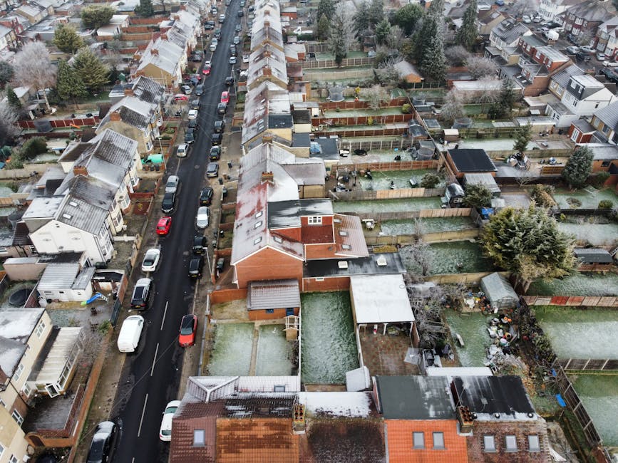 An aerial view of a suburban residential area showing a row of terraced houses with small, well-maintained gardens and driveways. The houses have brown tiled roofs, some with solar panels, and a mix of brick and painted exteriors. In the foreground, a large parking lot with marked spaces and multiple parked cars is visible adjacent to a single-story building. Surrounding the houses, there are trees, green lawns, and narrow pathways connecting the properties. The background features a busy street with additional commercial and residential blocks, including larger apartment buildings and a parking area filled with vehicles. The scene is captured in daylight with natural light illuminating the scene evenly, highlighting the residential and urban environment. The image exemplifies typical urban sprawl with a combination of private gardens and shared parking spaces, reflecting aspects of modern waste management and independent disposal practices, as often handled by local rubbish removal services, such as Waste Removal Dulwich.