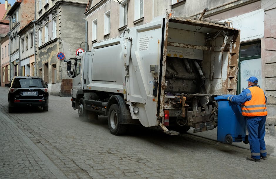 A street scene showing a white waste collection vehicle with an open rear compartment, which appears to be in the process of emptying or collecting rubbish. The vehicle is parked parallel to a row of older, multi-story buildings with weathered facades, some featuring peeling paint and brickwork. To the right, a worker dressed in a blue uniform and a high-visibility orange vest and hat is handling a blue wheeled bin, positioning it near the vehicle’s rear opening for waste transfer. Another dark-colored car is parked further along the cobblestone-paved street, which is narrow and typical of an urban residential area. The environment shows signs of urban life, with pavement markings and street signs visible, indicating parking restrictions. The lighting is neutral, suggesting an overcast day, and the scene reflects a typical private rubbish removal operation carried out by Waste Removal Dulwich as part of on-site waste management services aimed at responsible disposal of refuse in the local area.