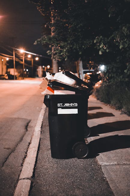 A black wheeled rubbish bin is positioned on the sidewalk at night, with its lid open and various waste materials visible inside, including paper and cardboard. The bin has a white label with the text 'ST. JOHN'S' printed on it, and it appears to be made of durable plastic with a smooth surface. The surrounding environment includes a dimly lit street with warm streetlights casting a glow, and trees with dark foliage casting shadows on the ground. In the background, blurred street elements such as streetlights, parked cars, and utility poles are visible, indicating a residential area. The scene suggests that the bin might be used for private waste disposal or collection services, aligning with independent rubbish removal options offered by waste management services like Waste Removal Dulwich. The image captures a typical urban setting where outdoor waste is temporarily stored for collection, highlighting the importance of proper waste handling and the role of local waste removal providers in managing refuse outside of municipal collection schedules.