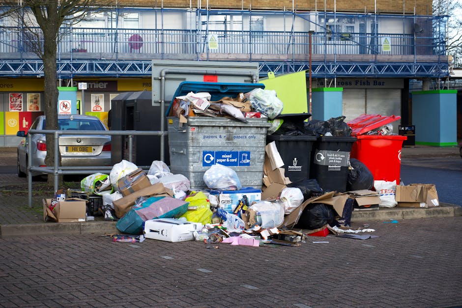 A street scene showing a white waste collection vehicle with an open rear compartment, which appears to be in the process of emptying or collecting rubbish. The vehicle is parked parallel to a row of older, multi-story buildings with weathered facades, some featuring peeling paint and brickwork. To the right, a worker dressed in a blue uniform and a high-visibility orange vest and hat is handling a blue wheeled bin, positioning it near the vehicle’s rear opening for waste transfer. Another dark-colored car is parked further along the cobblestone-paved street, which is narrow and typical of an urban residential area. The environment shows signs of urban life, with pavement markings and street signs visible, indicating parking restrictions. The lighting is neutral, suggesting an overcast day, and the scene reflects a typical private rubbish removal operation carried out by Waste Removal Dulwich as part of on-site waste management services aimed at responsible disposal of refuse in the local area.