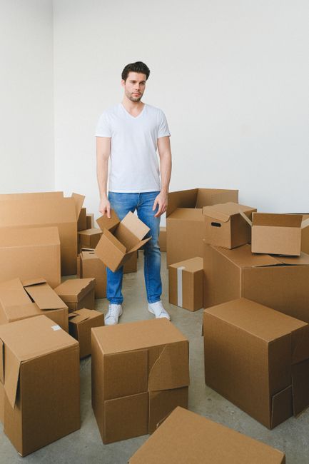 A young man with dark hair and light skin stands in a minimally decorated room with white walls, surrounded by numerous cardboard boxes of various sizes. The boxes are unsealed, some partially open or slightly crushed, and are arranged haphazardly on the floor and against the wall. The man is wearing a plain white T-shirt, blue jeans, and white sneakers, and he holds two smaller open boxes, one in each hand, with a neutral expression on his face. The environment suggests an on-site clearance or private waste handling scenario, typical for arrangements involving rubbish removal services such as Waste Removal Dulwich, highlighting the process of clearing clutter for estate clearances or similar projects.