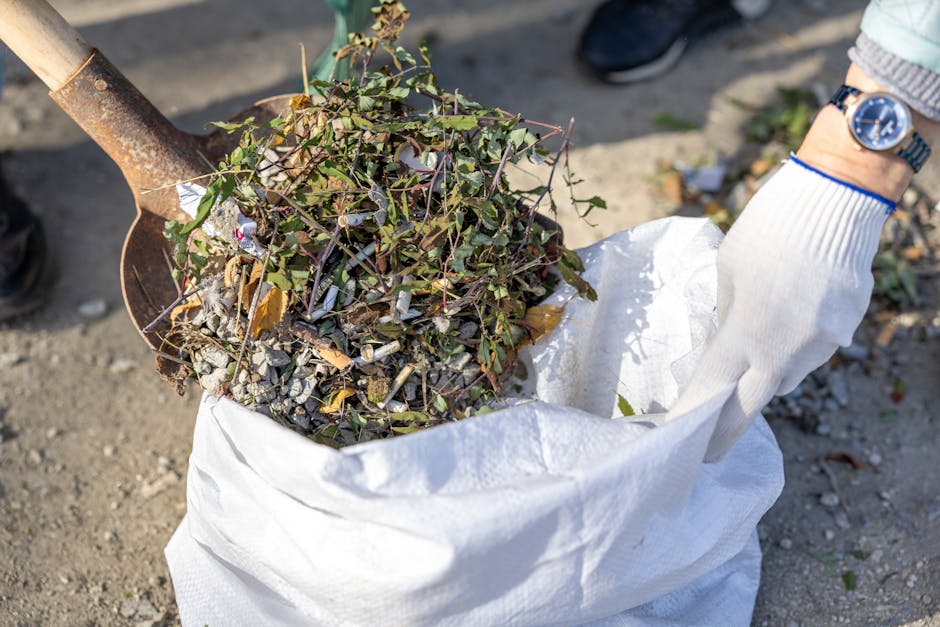 A close-up photograph of a pair of hands wearing white gloves holding a large white sack filled with garden debris and small branches with green and brown leaves. The sack appears to be made of woven polypropylene with a slightly wrinkled texture, and it is open at the top, revealing the organic waste inside. A rusty shovel with a wooden handle is partially visible, positioned behind the sack, indicating outdoor activity. The background shows a gravel or dirt surface with some scattered leaves and small debris, suggesting an outdoor setting for garden clearance or waste collection, related to independent rubbish removal services. The lighting is natural, casting subtle shadows on the ground and the objects in the scene, emphasizing the textures and materials involved in waste handling. This scene exemplifies on-site clearance activities often carried out by waste removal companies such as Waste Removal Dulwich, supporting private property waste management and alternative disposal options.