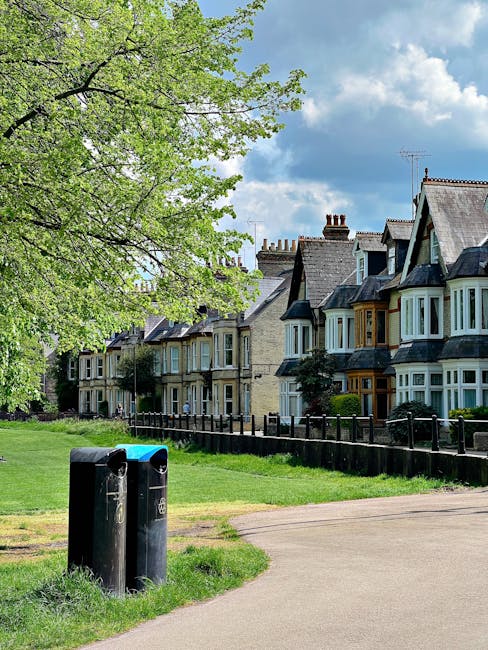 A residential street scene showing two black public recycling bins placed on a grassy verge near a paved footpath. Behind the bins, there's a row of traditional Victorian-style terraced houses with bay windows, brick facades, and chimneys, aligning along the street. To the left, a large tree with bright green fresh leaves extends its branches over the scene, offering partial shade. The sky above is mostly cloudy with patches of blue, creating diffuse natural light across the area. Visible behind the houses are additional rooftops and chimneys. The setting suggests an urban or suburban environment with nearby green space, indicative of a typical neighborhood that might alternatively use private waste collection services, which Waste Removal Dulwich can facilitate for on-site clearance or alternative rubbish disposal solutions.