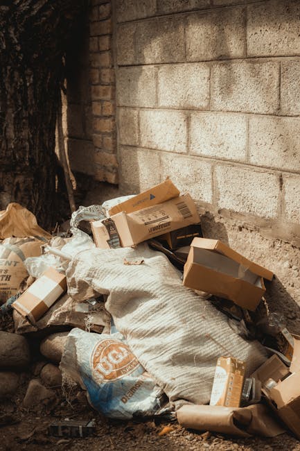 A collection of discarded cardboard boxes, some flattened and others partially assembled, with visible corrugated textures and brown and cardboard tones, piled against a gray brick wall outdoors. The boxes vary in size, with some leaning or resting on each other, and a large woven sack lies amidst the debris. The scene is set at the base of a rough brick and mortar wall, possibly part of an alleyway or garden boundary, with a dark tree trunk visible to the left. The natural outdoor lighting highlights the textures and colors of the packaging materials, emphasizing the presence of waste that may be targeted for private rubbish removal services, such as those offered by Waste Removal Dulwich. The cluttered arrangement and mixture of materials suggest an on-site clearance or an accumulation awaiting collection, reflecting typical rubbish removal scenarios in residential or commercial contexts.